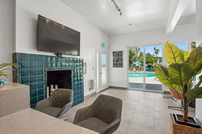 Modern lounge area with cozy chairs, a stylish tiled fireplace, and a large TV, leading to a vibrant pool view through glass doors.