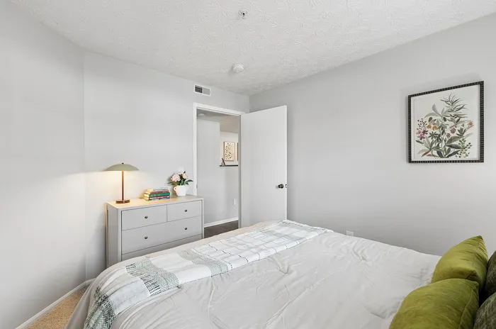 Tranquil bedroom with elegant decor, featuring a cozy bed with green accent pillows, a modern dresser with a stylish lamp, and a floral wall art piece.