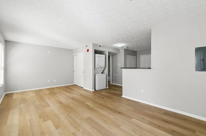 Bright open-concept living space with modern light wood flooring, featuring a compact stacked washer-dryer unit and ample natural light.