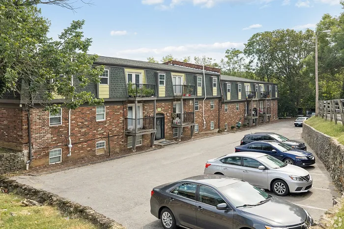 Charming brick apartment complex with modern balconies, surrounded by lush greenery and ample parking space.