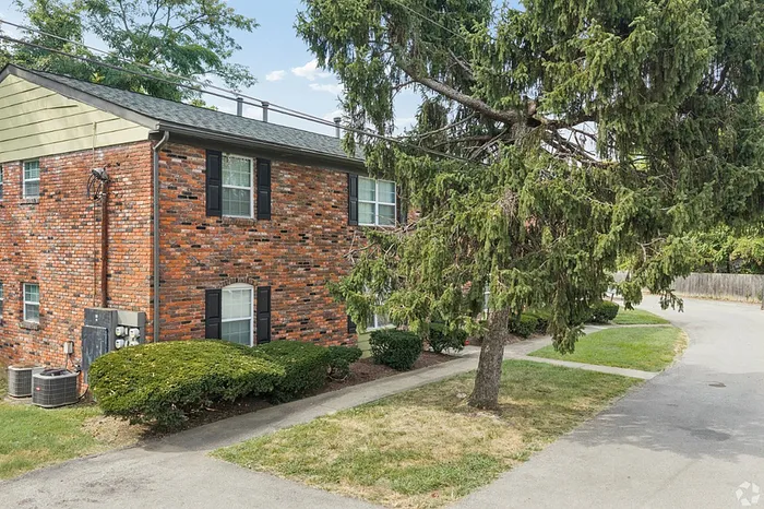 Charming brick apartment building with black shutters, nestled in a lush green setting, featuring a serene driveway and mature trees.
