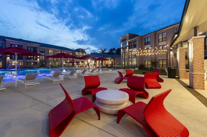 Luxury apartment complex courtyard at dusk with glowing pool, modern red lounge chairs, and string lights.
