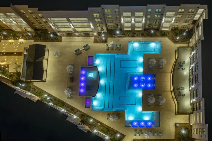 Stunning aerial view of a luxurious apartment complex with an illuminated swimming pool and inviting lounge areas, set against a modern building backdrop.