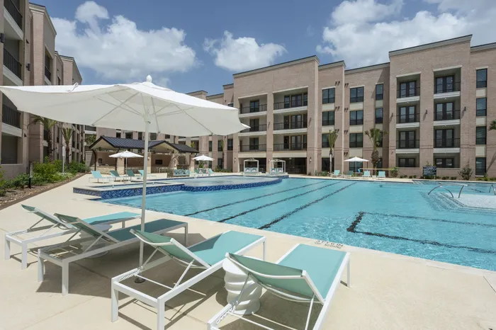 Luxurious apartment complex with a sparkling outdoor swimming pool, elegant poolside loungers, and shaded cabanas under clear blue skies.