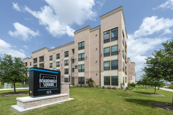 Modern brick apartment complex at Boardwalk Square with lush green lawns and expansive windows under a clear blue sky.