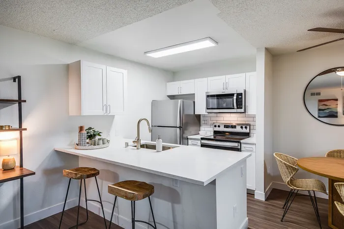 Modern kitchen with sleek white cabinetry, stainless steel appliances, and a cozy breakfast bar with stylish wooden stools, adjacent to a dining area with chic decor.