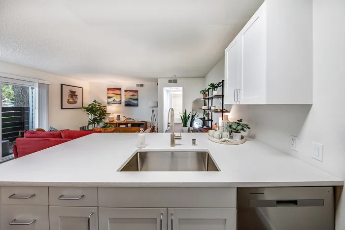 Modern open-concept kitchen with sleek countertops, stainless steel sink, and a view into a bright, inviting living room with vibrant decor and lush greenery.
