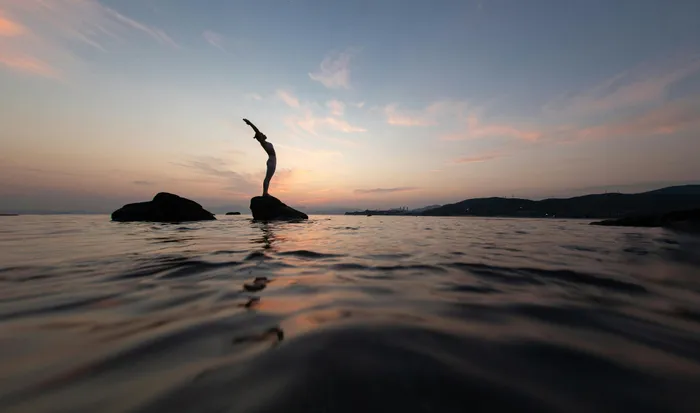 Serene silhouette of person stretching on seaside rocks at dusk, with peaceful ocean waves and vibrant sky backdrop.