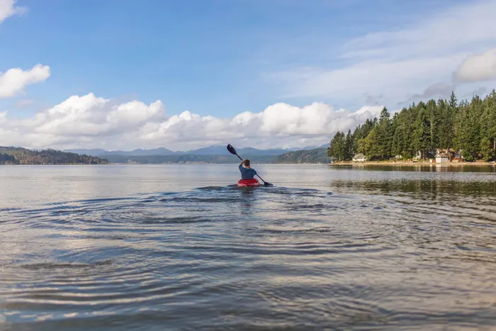 Serene lake view with person kayaking under a bright blue sky, surrounded by lush green forest and distant mountains.