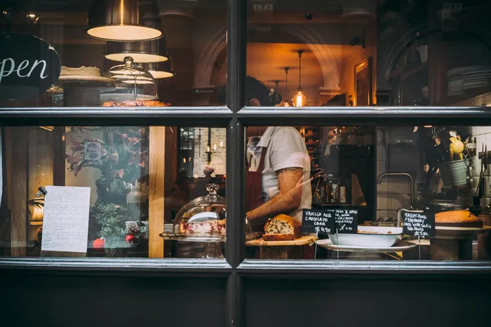 Cozy bakery interior with warm lighting, featuring displayed cakes and a person preparing desserts behind a glass window.