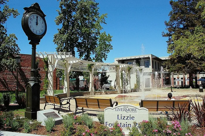 Charming park scene with elegant fountain, inviting benches, classic clock tower, and vibrant greenery under a clear blue sky.