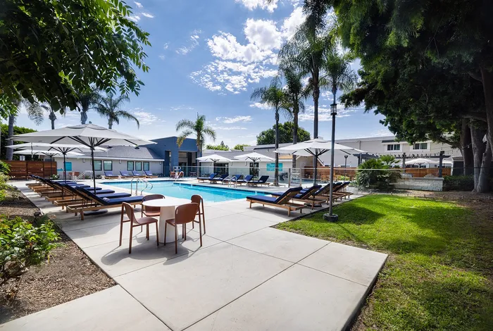 Elegant outdoor pool area with luxurious lounge chairs and umbrellas, surrounded by lush greenery under a vibrant blue sky.