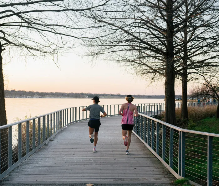 Two runners enjoying a scenic lakeside trail at sunset, surrounded by tranquil water and tall trees.