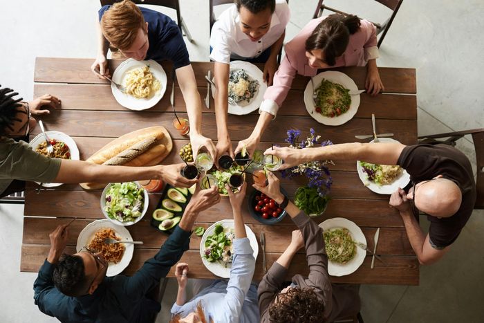 Top view of diverse people enjoying a vibrant meal, clinking glasses over a table filled with fresh salads, pasta, and bread.