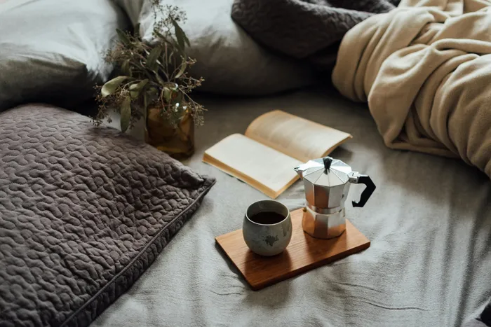 Cozy bedroom setting with plush pillows, a warm blanket, and a tray featuring a coffee maker and cup alongside an open book, creating a relaxing atmosphere.