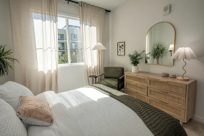 Sunlit bedroom featuring a cozy bed with white linens, elegant wooden dresser, and stylish green chair, embodying modern comfort and relaxation.