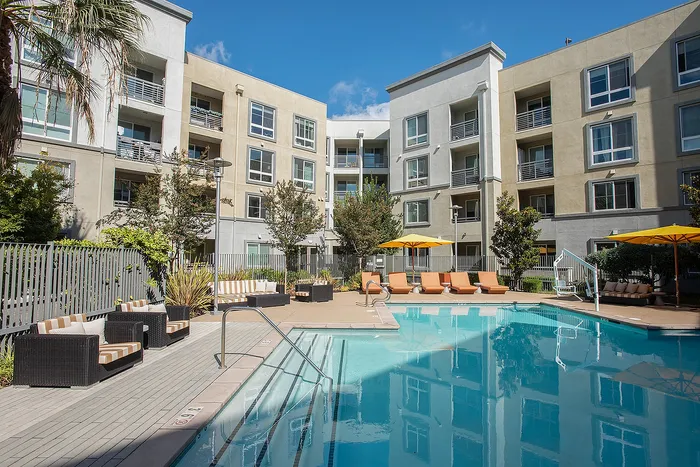 Modern apartment complex with serene outdoor pool area, surrounded by lush landscaping and stylish seating under vibrant umbrellas.