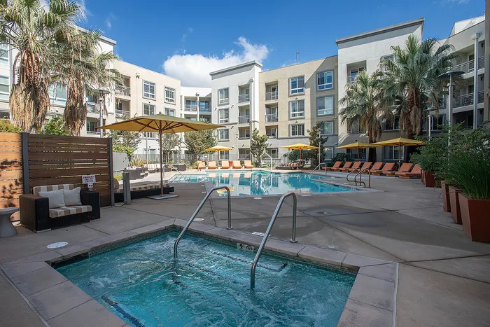 Luxurious apartment complex with a sparkling pool, cozy seating, and vibrant palm trees against a clear blue sky.