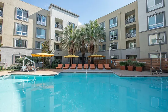 Modern apartment complex with a sparkling swimming pool, surrounded by lounge chairs and vibrant yellow umbrellas, offering a resort-like atmosphere.