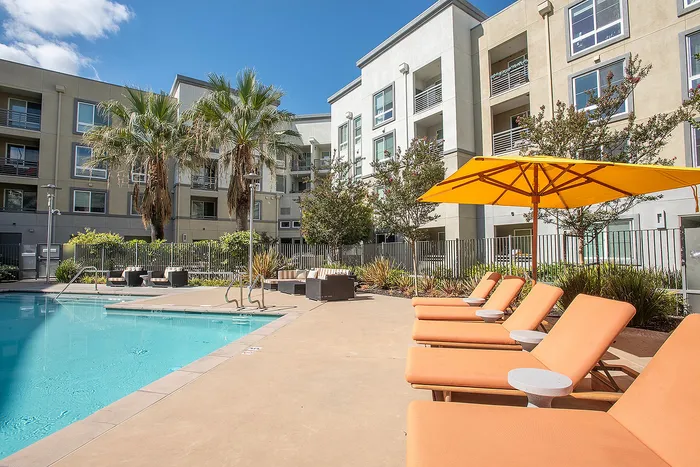 Inviting apartment complex featuring a sparkling pool, vibrant orange lounge chairs, and lush palm trees under a clear blue sky.