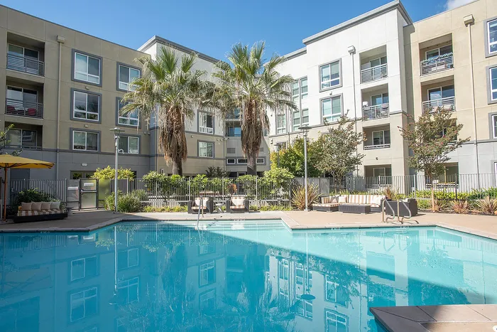 Inviting apartment complex featuring a sparkling pool, vibrant orange lounge chairs, and lush palm trees under a clear blue sky.
