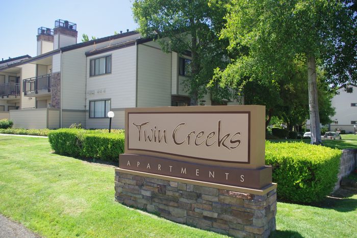 Twin Creeks Apartments entrance sign framed by green hedges and mature trees, with residential buildings in the background.