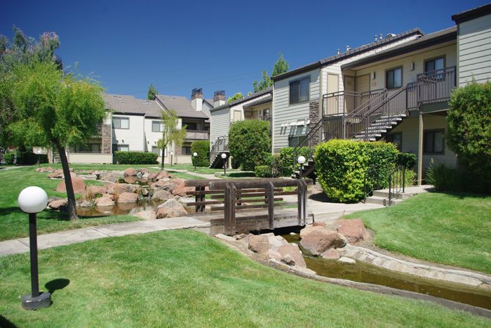 Twin Creeks residential buildings with a wooden footbridge crossing a rock-lined stream surrounded by green lawn.