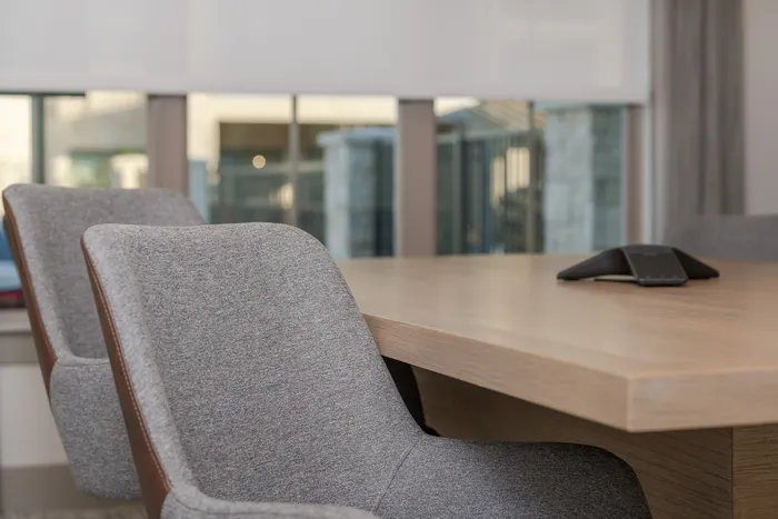 Modern conference room with sleek grey chairs and a polished wooden table, illuminated by natural light through large windows.