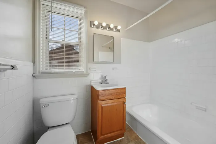 Well-lit modern bathroom featuring a clean white tiled bathtub, vanity with wooden cabinetry, and a large window.