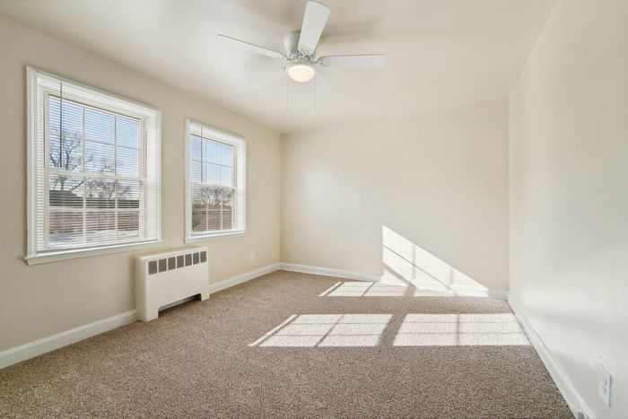 A sunlit, carpeted room with two large windows and a ceiling fan.