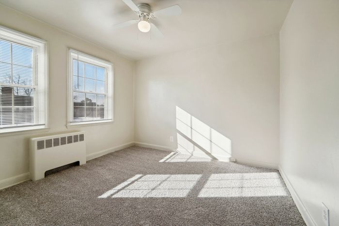 Bright empty room with beige carpet, large windows, and a ceiling fan.