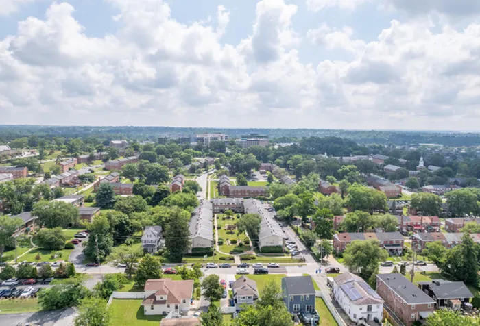 Charming aerial view of a sprawling suburban neighborhood with lush greenery, quaint houses, and a clear blue sky, perfect for tranquil living.