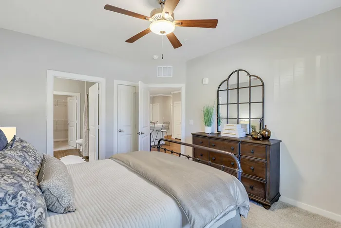 Bright and inviting bedroom featuring elegant bedding, a stylish ceiling fan, and a charming dresser with decorative accents.