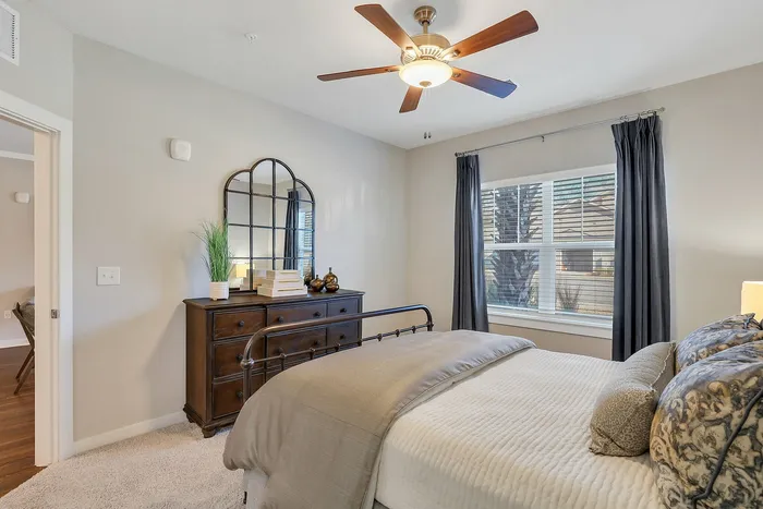 Cozy and elegant bedroom with a stylish ceiling fan, mirrored dresser, and large window offering ample natural light.