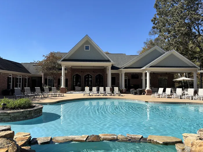 Inviting apartment complex with a pristine swimming pool, surrounded by stylish lounge chairs and lush trees under a clear blue sky.