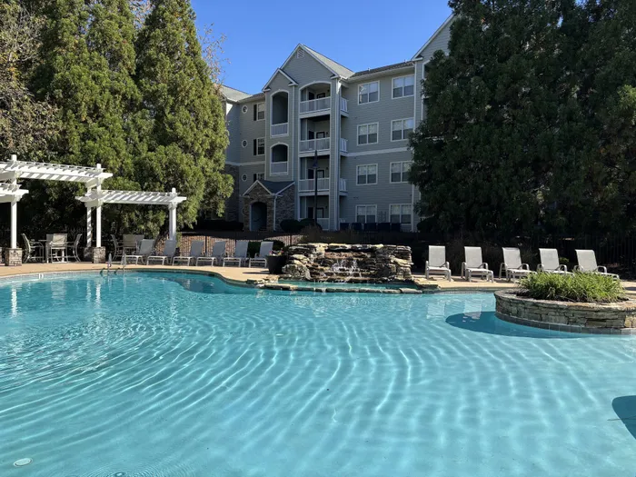Inviting apartment pool area with sun loungers and elegant pergola, surrounded by lush greenery and modern residential buildings.