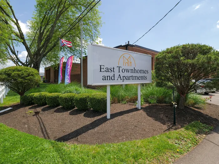 Inviting entrance sign for East Townhomes and Apartments surrounded by lush greenery and bright flags, capturing a warm and welcoming atmosphere.