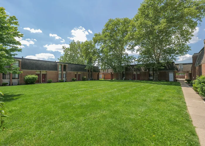 Charming courtyard view of brick apartment complex surrounded by lush greenery and mature trees under a clear blue sky.
