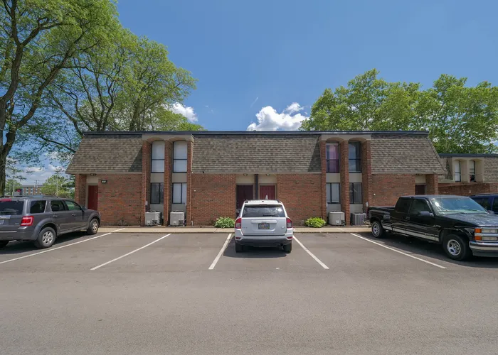 Charming brick apartment complex with ample parking, surrounded by lush trees and clear blue skies, creating an inviting living environment.