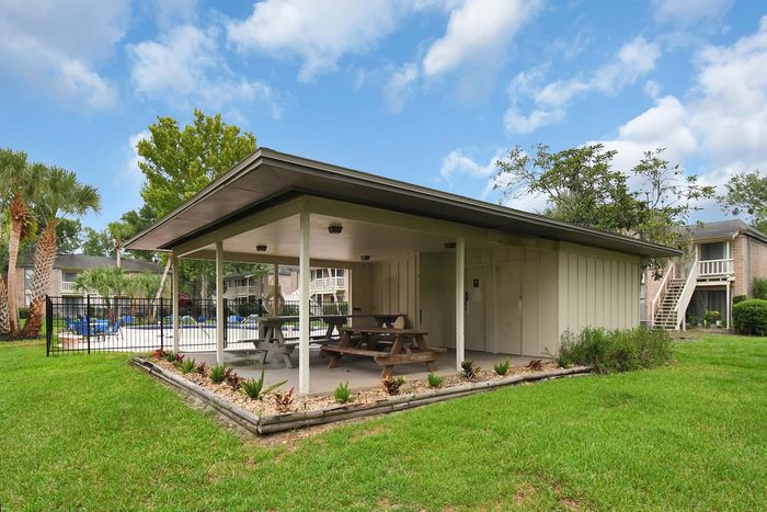 Charming outdoor pavilion with picnic tables set amidst lush greenery, adjacent to a sparkling pool and apartment buildings, perfect for outdoor gatherings.