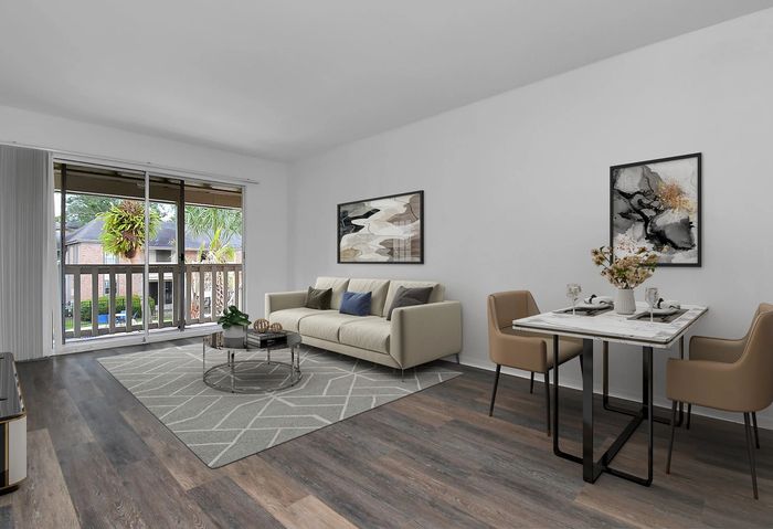 Modern living and dining area with sleek wood flooring, comfy beige sofa, elegant artwork, and ample natural light from large windows overlooking lush greenery.