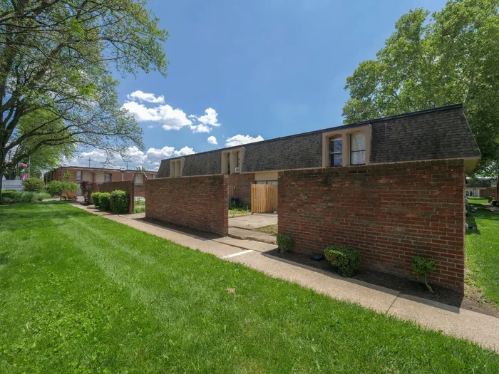 Charming brick apartment complex with beautifully landscaped walkways and lush green lawns under a clear blue sky.