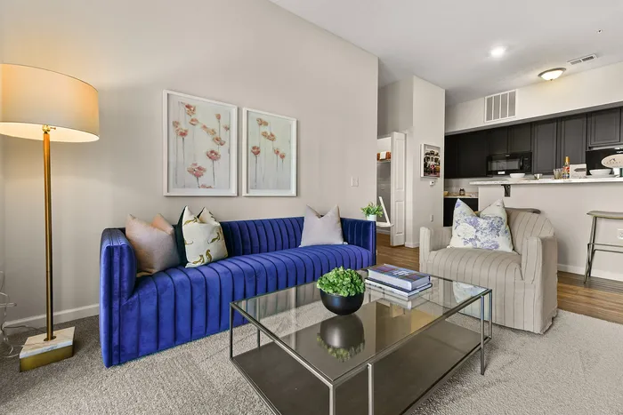 Chic living room showcasing a vibrant blue sofa, elegant armchair, and a modern glass coffee table against a backdrop of stylish decor and an inviting open kitchen.