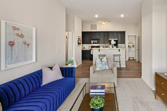 Modern open-plan living area with a vibrant blue sofa, elegant armchairs, and a sleek kitchen in the background featuring a breakfast bar with stools.