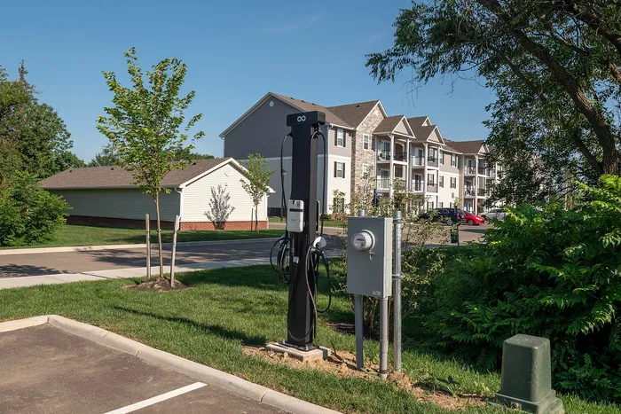 Modern apartment complex with electric vehicle charging station, surrounded by lush greenery and clear skies.
