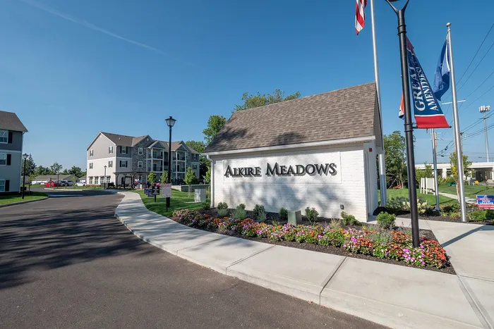 Charming Alkire Meadows entrance with vibrant flowerbeds and modern apartment buildings under a clear blue sky.