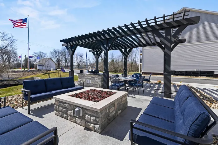 Inviting outdoor patio with stylish pergola, cozy seating around a central stone fire pit, and dining area under blue skies.