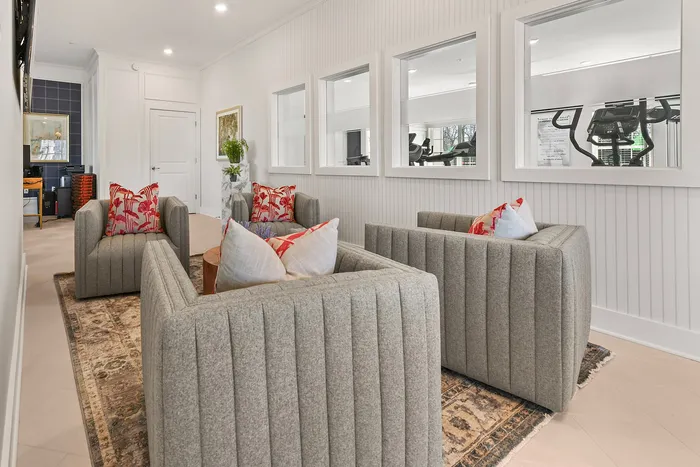 Inviting lounge area featuring plush grey armchairs with vibrant pillows, elegant wall decor, and a view into the gym through stylish windows.