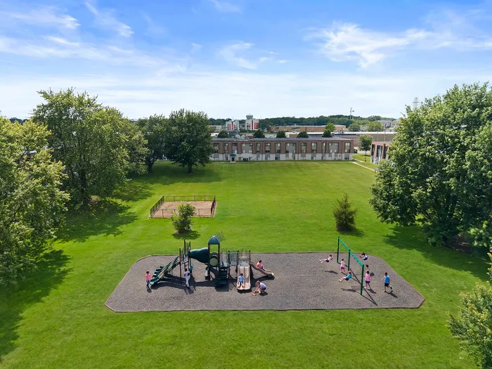 Expansive green space featuring a vibrant playground with lively children, surrounded by lush trees and modern apartment buildings in the background.