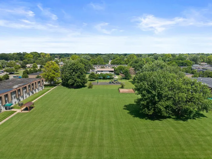 Aerial view of a lush green park with tree-lined walkways and adjacent residential buildings under a clear blue sky.